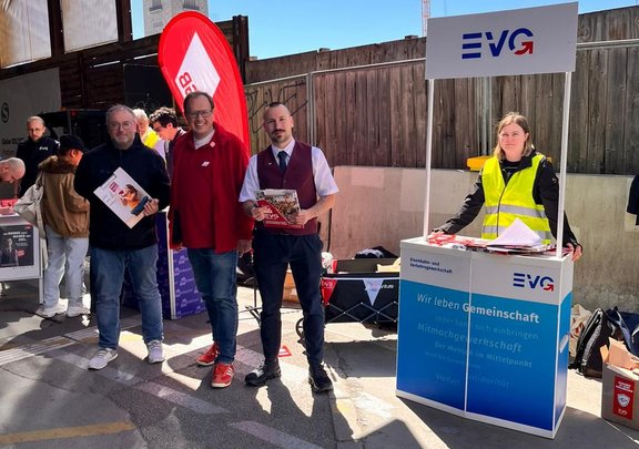 Mehrere Personen stehen im Freien an einem Informationsstand. Rechts steht ein mobiler Stand mit der Aufschrift „EVG“ und „Wir leben Gemeinschaft“. Auf dem Stand liegen Flyer und Unterlagen. Eine Person trägt eine gelbe Warnweste hinter dem Stand. Links davon stehen drei weitere Personen nebeneinander, zwei halten Broschüren in den Händen. Im Hintergrund sind Holzverschläge, Banner und weitere Stände zu sehen.
