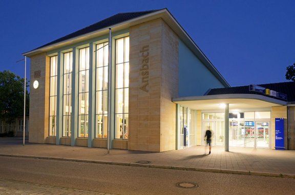 Beleuchtetes Bahnhofsgebäude in der Dämmerung mit heller Natursteinfassade, hohen schmalen Fensterflächen und dem sichtbar eingravierten Schriftzug „Ansbach“ an einer Gebäudeecke. Links an der Fassade sind ein DB-Logo und eine beleuchtete Uhr zu sehen. Rechts befindet sich ein überdachter Eingangsbereich mit Glasfronten und automatischen Türen. Vor dem Gebäude verläuft eine gepflasterte Fläche mit Fahrbahnrand; nahe dem Eingang ist eine verschwommen dargestellte gehende Person erkennbar. Im Hintergrund sind Bäume und weitere Gebäudeteile sichtbar.