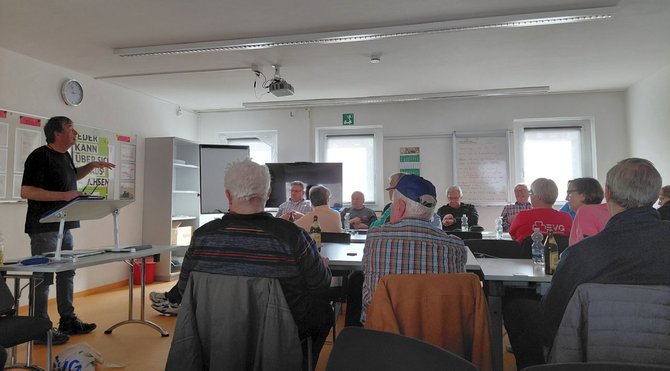 Eine Gruppe von Personen sitzt in einem Seminarraum an Tischen, die in U‑Form angeordnet sind. Im Vordergrund sind mehrere Personen von hinten zu sehen. Vorn links steht eine Person an einem Rednerpult. An den Wänden hängen Whiteboards und Flipcharts mit Notizen. In der Mitte des Raums steht ein großer Bildschirm. Auf den Tischen stehen Getränke und verschiedene Materialien.