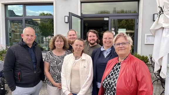 Sieben Personen stehen vor dem Eingang eines Gebäudes mit großen Fenstern und geöffneter Tür. Die Personen sind in zwei Reihen angeordnet. Im Vordergrund sind verschiedene Jacken, Oberteile und Brillen sichtbar. Rechts und links am Gebäude befinden sich Außenleuchten, am rechten Bildrand steht ein geschlossener Sonnenschirm. Vor dem Gebäude stehen Tische und Stühle auf gepflastertem Boden.