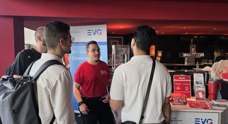 In einem Innenraum mit einem Informationsstand und einem Banner mit der Aufschrift „EVG“ stehen mehrere Personen im Gespräch. Auf dem Tresen liegen Broschüren und Informationsmaterial. Ein Schild mit der Aufschrift „Tag der Nachwuchskräfte 17.09.26 11–18 Uhr“ ist sichtbar. Im Hintergrund befinden sich weitere Einrichtungsgegenstände und eine Getränkestation.