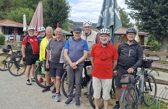 Acht Personen stehen mit Fahrrädern auf einem Weg vor einem Gebäude mit Außengastronomie. Mehrere Personen tragen Fahrradhelme und Radbekleidung. Im Hintergrund sind Sonnenschirme, Bäume und bewaldete Hügel unter einem bewölkten Himmel zu sehen. Einige Fahrräder stehen neben der Gruppe, darunter ein Fahrrad im Vordergrund rechts.