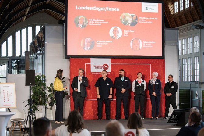 Mehrere Personen stehen auf einer Bühne vor einem roten Hintergrund mit einer großen Leinwand darüber. Auf der Leinwand steht „Landessieger/innen“ sowie mehrere Namen mit kleinen Porträtbildern. Die Personen auf der Bühne stehen in einer Reihe, einige tragen Namensschilder. Links steht eine Person mit Mikrofon, rechts sind weitere Personen nebeneinander aufgestellt. Im Vordergrund ist Publikum zu sehen.