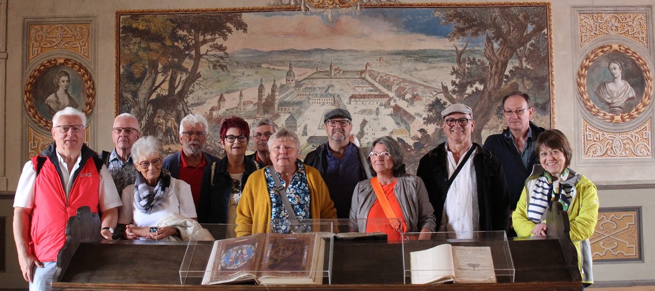 Eine Gruppe von etwa zwölf Personen steht in einem Innenraum vor einer Wand mit großem Wandgemälde und mehreren ausgestellten alten Büchern. Die Personen stehen dicht nebeneinander hinter einer langen Vitrine aus Holz mit Glasabdeckungen. In der Vitrine liegen zwei aufgeschlagene, sehr alte Bücher mit verzierten Seiten. Einige der Personen tragen Jacken oder Westen, eine Person trägt eine rote Weste mit sichtbarer Aufschrift „EVG“. Mehrere tragen Brillen oder haben kleine Taschen umgehängt. Alle blicken in Richtung Kamera.