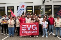 Eine große Gruppe von Personen steht vor einem Gebäude mit mehreren Fenstern und einer geöffneten Tür. In der Mitte hält die Gruppe ein rotes Banner mit der weißen Aufschrift „EVG JUGEND NRW“. Links und rechts werden Fahnen gehalten, darunter eine weiße Fahne mit einem Logo und eine rote Fahne. Die Personen tragen überwiegend helle Oberteile und rote Namensbänder um den Hals. Im Hintergrund sind weitere Personen auf einer Treppe vor dem Eingang zu sehen. Der Boden ist gepflastert, und es handelt sich um eine Aufnahme im Freien bei Tageslicht.
