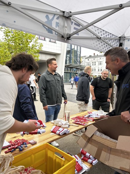 Blick von unter einem Pavillon auf einen Informationsstand mit Holztischen. Auf den Tischen liegen rote Flyer, kleine verpackte Gegenstände, weiße Textilien und Stapel von Karten. Rechts steht ein großer geöffneter Karton. Mehrere Personen befinden sich am Stand oder auf dem Platz dahinter. Im Hintergrund sind Gebäude, Bäume und Passanten sichtbar.