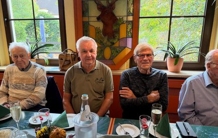 Vier Personen sitzen an einem gedeckten Tisch vor großen Fenstern. Auf der Fensterbank stehen mehrere Topfpflanzen. In der Mitte des Fensters befindet sich ein farbiges Glasbild mit einem Hirschmotiv. Auf dem Tisch stehen Teller, Gläser, gefaltete Servietten, eine Wasserflasche in einem Kühler und eine kleine Herbstdekoration.