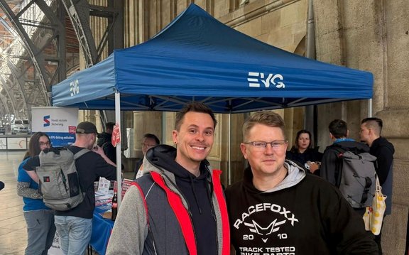 Unter einem blauen Pavillon mit EVG Logo stehen mehrere Personen in einer Bahnhofshalle. Im Vordergrund posieren zwei Personen für ein Foto. Im Hintergrund befindet sich ein Infostand mit Roll Up Banner und weiteren Personen, die sich dort aufhalten. Das Bild zeigt Säulen und eine überdachte Bahnhofskonstruktion.