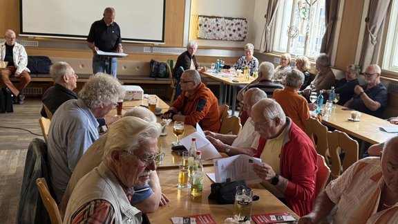 Großer Innenraum mit Holzboden, hellen Wänden und Fenstern an der rechten Seite. Mehrere lange Holztische stehen parallel, an denen viele Personen auf Stühlen sitzen. Auf den Tischen liegen Papierunterlagen, Mappen, Stifte und rote Flyer; außerdem stehen Gläser mit Getränken, Flaschen mit Wasser und Biergläser mit aufgedrucktem Schriftzug. Im Hintergrund befindet sich eine Leinwand; davor steht eine Person mit Papieren in den Händen. Weitere Personen sitzen an einem Tisch nahe der Leinwand. An den Wänden hängen Vorhänge und ein dekoratives Wandobjekt. Ein Beamer steht auf einem Tisch im vorderen Bereich. Die Szene wirkt wie eine Versammlung oder Besprechung in einem Gastraum.
