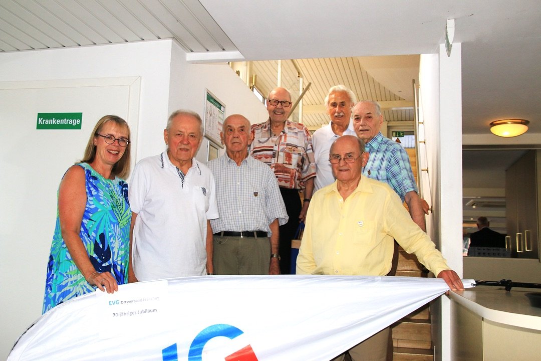 Sieben Personen stehen im Innenbereich eines Schiffes vor einem Treppenaufgang hinter einer EVG-Fahne und posieren für ein Gruppenfoto. Die Gruppe befindet sich in einem hellen Raum vor einer Treppe mit Handläufen. Im Vordergrund ist eine weiße EVG-Fahne zu sehen. Auf der Fahne ist ein Schild mit der Aufschrift „EVG Ortsverband Frankfurt“ und „70-jähriges Jubiläum“ angebracht. Die Personen stehen teils vor der Treppe, teils auf den ersten Stufen.