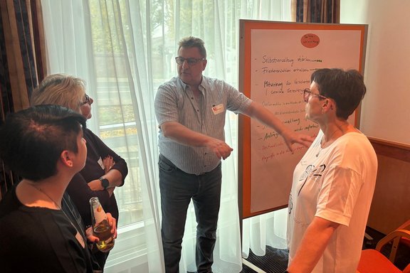 Vier Personen stehen in einem Seminar- oder Gruppenraum um ein Flipchart, während eine Person vor dem Flipchart mit den Armen auf die Tafel und die Gruppe zeigt. Das Foto zeigt eine Arbeitssituation in kleiner Runde. Drei Personen stehen dem Flipchart zugewandt und hören zu, während die Person in der Mitte vor der Tafel steht und auf Inhalte verweist. Das Flipchart enthält handschriftliche Stichpunkte. Lesbar sind unter anderem „Selbstverwaltung stärken“, „Friedenswahlen erhalten“ sowie Hinweise zur Mitwirkung der Versicherten und zur sozialen Gerechtigkeit.