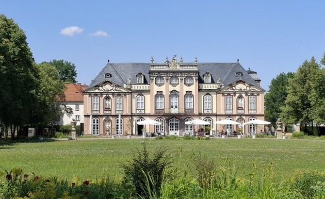 Symmetrisches, repräsentatives Gebäude mit drei Geschossen, heller Fassade und dunklem Mansarddach, mittig auf einer großen grünen Rasenfläche. Vor dem Gebäude stehen mehrere weiße Sonnenschirme und Sitzbereiche. Bäume rahmen die Szene links und rechts ein, darüber ein klarer blauer Himmel mit wenigen kleinen Wolken.