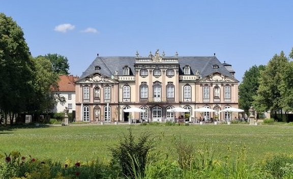 Symmetrisches, repräsentatives Gebäude mit drei Geschossen, heller Fassade und dunklem Mansarddach, mittig auf einer großen grünen Rasenfläche. Vor dem Gebäude stehen mehrere weiße Sonnenschirme und Sitzbereiche. Bäume rahmen die Szene links und rechts ein, darüber ein klarer blauer Himmel mit wenigen kleinen Wolken.