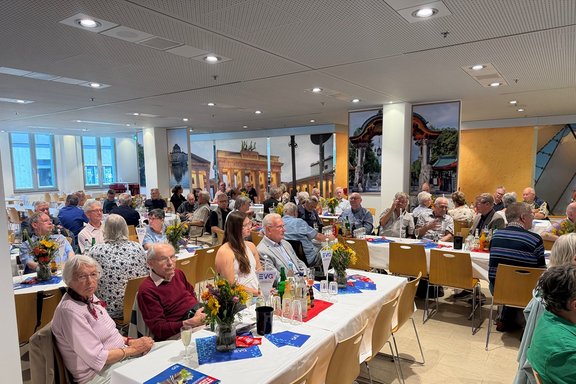 Zahlreiche Menschen sitzen an festlich gedeckten Tischen in einem Veranstaltungsraum und nehmen an einer gemeinsamen Feier oder Versammlung teil. Der Raum ist mit mehreren langen Tischreihen ausgestattet, die überwiegend besetzt sind. Die Tische sind mit weißen Tischdecken, Blumensträußen, Getränken, Gläsern und Informationsmaterialien dekoriert. Viele Personen sitzen miteinander an den Tischen und richten den Blick in verschiedene Richtungen des Raumes. Im Vordergrund sind mehrere gedeckte Tische mit Blumenschmuck und kleinen Tischaufstellern zu sehen.