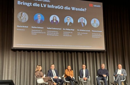 Podiumsdiskussion auf einer Bühne mit sechs sitzenden Personen. Über der Bühne zeigt eine große Projektionsleinwand den Titel „Bringt die LV InfraGO die Wende?“. Darunter sind sechs Porträtbilder mit Namen und Funktionsbezeichnungen abgebildet. Rechts oben auf der Leinwand ist das Logo „DB InfraGO“ sichtbar. Vor der Leinwand stehen sechs Stühle in einer Reihe auf der Bühne vor einem grauen Vorhang.