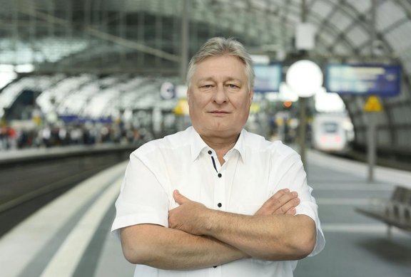 Person in einem weißen Kurzarmhemd steht mit verschränkten Armen auf einem Bahnsteig. Im Hintergrund sind unscharf Gleise, ein Zug, Bahnsteiganzeigen und die gewölbte Dachkonstruktion eines großen Bahnhofs zu sehen.