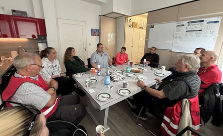 Neun Personen sitzen in einem Besprechungsraum um einen rechteckigen Tisch. Auf dem Tisch stehen Teller, Tassen, Gläser, Wasserflaschen und mehrere Kuchen. Im Hintergrund befinden sich eine Küchenzeile, ein Whiteboard, Schränke und eine geöffnete Tür.