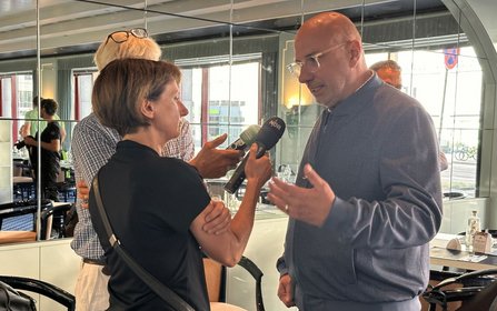 Eine Person steht in einem Innenraum und spricht in mehrere Mikrofone, die von zwei anderen Personen gehalten werden. Hinter den Personen befinden sich Spiegelwände, Fenster und mehrere Stühle. Auf einem Mikrofon ist die Aufschrift „NDR“ sichtbar.