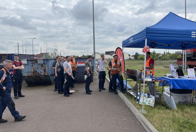 Eine größere Gruppe von Personen steht auf einem Gelände neben einem blauen „EVG“-Faltzelt. Mehrere Personen tragen orange Warnwesten, andere Arbeitskleidung. Rechts neben dem Zelt stehen Tische mit Getränken, Bechern und Materialien; daneben ein Grill. Links im Hintergrund befinden sich große Container, weiter hinten Gleise, Gebäude und Grünflächen.