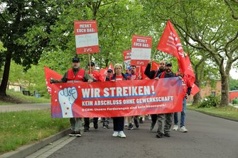 Demonstrationszug auf einer von Bäumen gesäumten Straße. Vorne wird ein großes rotes Banner mit der Aufschrift „WIR STREIKEN! KEIN ABSCHLUSS OHNE GEWERKSCHAFT“ getragen. Dahinter sind zahlreiche rote EVG-Fahnen und mehrere Schilder mit der Überschrift „MERKT EUCH DAS:“ zu sehen.