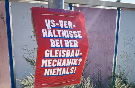 otes Schild an einer Haltestelle oder Werbefläche mit der Aufschrift: „US-VER-HÄLTNISSE BEI DER GLEISBAU-MECHANIK? NIEMALS!“. Hinter dem Schild befinden sich helle Wandflächen mit Graffiti und wild wachsende Pflanzen am Boden.