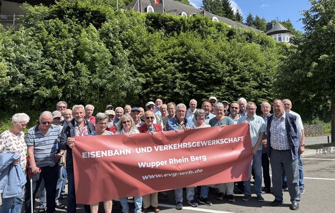 Eine große Gruppe von Menschen steht im Freien vor einer dichten grünen Böschung mit Bäumen und Sträuchern. Im Hintergrund ist ein Gebäude mit grauem Dach und mehreren Dachfenstern sowie eine runde Turmstruktur sichtbar. Die Gruppe hält ein großes rotes Banner mit weißer Aufschrift: „EISENBAHN UND VERKEHRSGEWERKSCHAFT“ sowie darunter „Wupper Rhein Berg“ und „www.evg-wrb.de“. Die Menschen stehen dicht nebeneinander und halten das Banner über die gesamte Breite. Einige tragen Sonnenbrillen, Hüte oder Kappen.