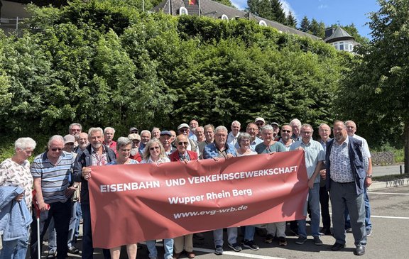 Eine große Gruppe von Menschen steht im Freien vor einer dichten grünen Böschung mit Bäumen und Sträuchern. Im Hintergrund ist ein Gebäude mit grauem Dach und mehreren Dachfenstern sowie eine runde Turmstruktur sichtbar. Die Gruppe hält ein großes rotes Banner mit weißer Aufschrift: „EISENBAHN UND VERKEHRSGEWERKSCHAFT“ sowie darunter „Wupper Rhein Berg“ und „www.evg-wrb.de“. Die Menschen stehen dicht nebeneinander und halten das Banner über die gesamte Breite. Einige tragen Sonnenbrillen, Hüte oder Kappen.