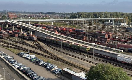 Weitwinkelansicht eines großen Güterbahnhofs mit zahlreichen Gleisen, abgestellten Güterwagen, Straßenbrücken und Oberleitungsanlagen. Im Vordergrund befinden sich ein Parkplatz mit mehreren Fahrzeugen sowie kleinere Gebäude. Im Hintergrund erstrecken sich weitere Gleisanlagen und Baumreihen unter bewölktem Himmel.