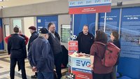 Mehrere Personen stehen in einer Bahnhofshalle an einem Infostand der EVG. Der Stand zeigt Schilder und Materialien mit dem EVG‑Logo und Texten. Auf dem Tresen liegen Flyer, kleine Gegenstände und ein aufgestelltes Schild. Hinter dem Stand befinden sich blau gestaltete Informationstafeln. Links im Hintergrund sind DB‑Hinweisschilder und Türen zu sehen.