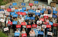 Große Gruppe von Menschen steht im Freien auf einer gepflasterten Fläche und hält vor sich oder über den Kopf zahlreiche rechteckige Schilder in den Farben Blau, Rot und Weiß. Auf den Schildern sind kurze Slogans in deutscher Sprache zu lesen, jeweils mit „EVG“ als Absender. Einige Personen halten zusätzlich eine blaue Fahne mit weißer Beschriftung. Im Hintergrund sind Pflanzen, ein Gebäude mit heller Fassade und gestapelte Holzscheite zu sehen. Die Personen sind unterschiedlich gekleidet, stehen dicht beieinander und blicken überwiegend zur Kamera.