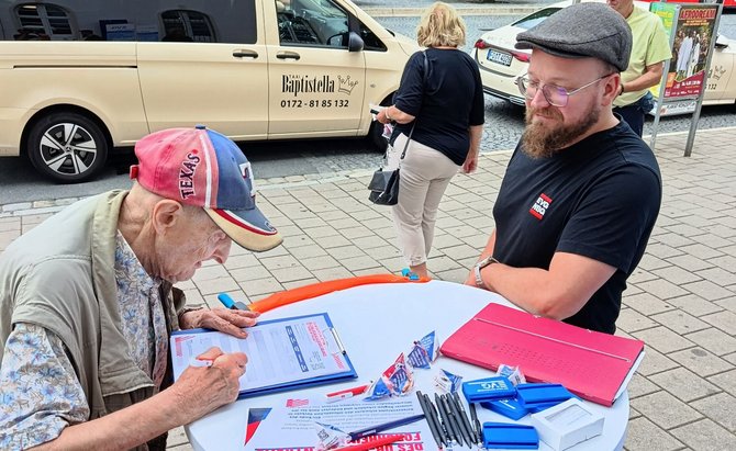 Ein älterer Mann mit Baseballkappe unterschreibt ein Formular auf einem Klemmbrett an einem Stehtisch, gegenüber steht ein Mann mit Mütze; auf dem Tisch liegen Flyer, Stifte und kleine Werbeartikel, im Hintergrund sind ein Taxi und Passanten zu sehen.