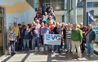 Große Gruppe von etwa 50 Personen in einem hellen Innenraum eines Gebäudes, angeordnet auf einer Treppe und dem unteren Podest. Im Vordergrund stehen mehrere Personen dicht nebeneinander, dahinter weitere Personen stufenweise nach oben. Die Personen tragen unterschiedliche Freizeit- und Alltagskleidung in verschiedenen Farben. In der Mitte der vorderen Reihe halten zwei Personen ein rechteckiges Banner mit der Aufschrift „EVG Eisenbahn- und Verkehrsgewerkschaft“ in blauer Schrift auf hellem Hintergrund. Der Raum hat große Fensterflächen, Glasgeländer an der Treppe, helle Wände und einen sichtbaren Eingangsbereich mit Türen. Tageslicht fällt von der Seite in den Raum.
