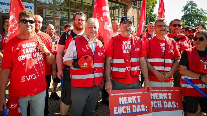 Gruppe von Personen vor einem Gebäude mit großen Fenstern. Viele tragen rote Westen und rote T-Shirts mit EVG-Logo sowie rote Fahnen. Im Vordergrund halten mehrere Personen rote Schilder mit der Aufschrift „MERKT EUCH DAS:“. Auf einem T-Shirt steht sichtbar „OHNE TARIF? OHNE UNS!“.