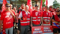 Gruppe von Personen vor einem Gebäude mit großen Fenstern. Viele tragen rote Westen und rote T-Shirts mit EVG-Logo sowie rote Fahnen. Im Vordergrund halten mehrere Personen rote Schilder mit der Aufschrift „MERKT EUCH DAS:“. Auf einem T-Shirt steht sichtbar „OHNE TARIF? OHNE UNS!“.