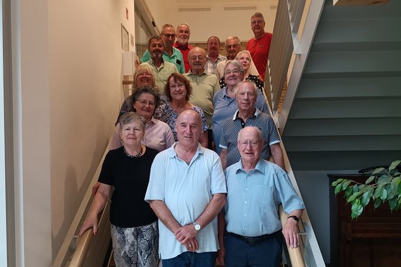 Eine Gruppe von 15 Personen steht auf einer breiten Innentreppe und blickt in die Kamera. Die Personen sind auf mehreren Stufen einer hellen Treppe angeordnet. Im Vordergrund stehen drei Personen auf den unteren Stufen, dahinter folgen weitere Personen bis zum oberen Treppenabsatz. Die Gruppe nimmt die gesamte Breite der Treppe ein.