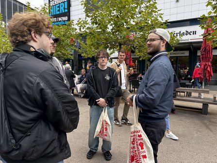 Außenbereich auf einem Platz mit Bäumen und Sitzgelegenheiten. Mehrere Personen stehen in einer Gesprächsgruppe. Einige tragen helle Stofftaschen mit rotem Aufdruck. Im Hintergrund sind Geschäfte, Passanten, Sitzbänke und ein Werbeschild an einem Gebäude zu sehen. Der Himmel ist bewölkt.