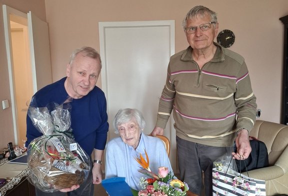 Drei Personen stehen bzw. sitzen in einem Zimmer. In der Mitte sitzt eine ältere Person an einem Tisch mit einem Blumenstrauß. Die Person links hält einen Geschenkekorb in Folie verpackt. Die Person rechts hält eine gestreifte Geschenktüte. Im Hintergrund sind eine geöffnete Tür, eine Wanduhr und ein Teil eines Sofas zu sehen.