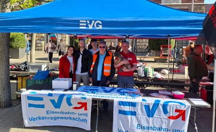 Unter einem blauen Pavillon mit der Aufschrift „EVG“ steht ein Tisch mit mehreren ausgelegten Bannern und Materialien. Auf den Bannern ist der Text „EVG Eisenbahn- und Verkehrsgewerkschaft“ gut lesbar. Hinter dem Tisch stehen fünf Personen nebeneinander; eine Person trägt eine orangefarbene Warnweste, eine andere ein rotes T‑Shirt mit EVG‑Logo und hält ein kleines Tier im Arm. Auf dem Tisch liegen Flyer, Stoffbänder und verschiedene Gegenstände; daneben stehen Kisten und Taschen. Im Hintergrund sind weitere Pavillons, Passanten, Bäume und Gebäude einer Innenstadt zu sehen.