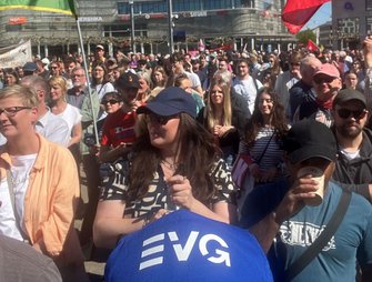 Große Menschenmenge auf einem Platz bei Tageslicht. Im Vordergrund sind dicht stehende Personen zu sehen, einige halten Fahnen oder Schilder. Eine blaue Kappe mit der Aufschrift „EVG“ ist im unteren Bildbereich sichtbar. Im Hintergrund stehen Gebäude mit gut lesbaren Geschäftslogos „SATURN“ und „BERSHKA“. Mehrere Fahnen in Rot sowie eine grün-gelbe Fahne ragen über die Menge.