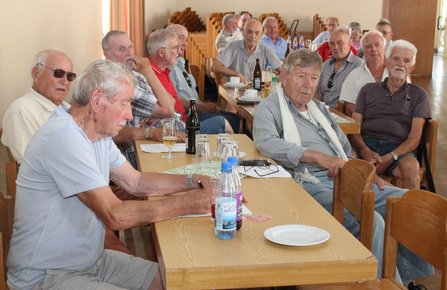 Mehrere ältere Männer sitzen an Tischen in einem Raum und schauen in Richtung Kamera. Auf den Tischen stehen Gläser, Flaschen und Unterlagen. Im Hintergrund sind gestapelte Stühle zu sehen.