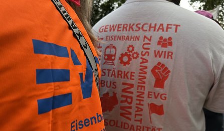Nahaufnahme einer orangefarbenen Warnweste mit blauem EVG-Logo im linken Bildbereich. Daneben ist der Rücken einer Person mit weißem T‑Shirt zu sehen. Auf dem T‑Shirt stehen rote Wörter und Symbole, darunter „GEWERKSCHAFT“, „WIR“, „EVG“, „ZUSAMMENHALT“, „SOLIDARITÄT“, eine Faust, ein Zug, Zahnräder und eine Fahne. Im Hintergrund sind weitere Personen und Bäume erkennbar.