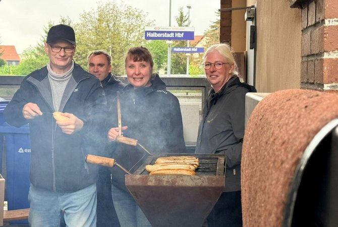 Mehrere Personen stehen im Freien an einem Grill. Auf dem Grill liegen mehrere Bratwürste, Rauch steigt auf. Eine Person hält eine Zange, eine andere ein Brötchen. Im Hintergrund ist ein Bahnhofsschild mit der Aufschrift „Halberstadt Hbf“ zu sehen.