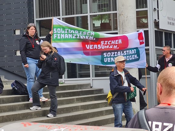 Mehrere Personen stehen auf einer Treppe vor einem Gebäude und halten ein großes Banner mit gewerkschaftlichen Forderungen und dem Logo von ver.di. Im Mittelpunkt des Bildes steht ein großes, quer gehaltenes Banner, das an zwei Seiten von Personen festgehalten wird. Auf dem Banner stehen farbige Abschnitte mit den Texten: „SOLIDARISCH FINANZIEREN.“, „SICHER VERSORGEN.“ und „SOZIALSTAAT VERTEIDIGEN.“ Rechts oben ist ein rotes Feld mit dem Logo „ver.di“ zu sehen. Mehrere Personen tragen dunkle Jacken und teilweise rote Bänder oder Schilder um den Hals. Eine Person links oben hält das Banner an der Treppe, während eine weitere Person daneben auf ein Mobiltelefon schaut. Im Vordergrund steht eine Person mit einer Mütze und einer Tasche, die leicht seitlich zur Kamera steht.