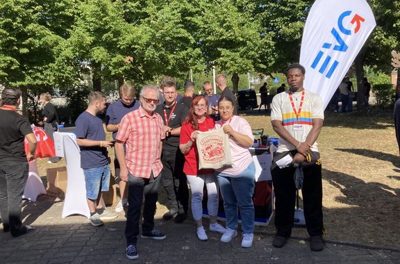 Mehrere Personen stehen an einem Informationsstand im Freien unter Bäumen. Im Vordergrund posieren vier Personen vor einem Tisch mit Informationsmaterial. Eine Person hält einen Stoffbeutel mit rotem Aufdruck. Rechts steht eine weiße Fahne mit blau-rotem Logo. Weitere Personen befinden sich im Hintergrund zwischen Tischen und Ausstellungsständen.