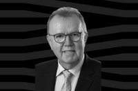 Ein Schwarz-Weiß-Porträt zeigt eine Person mit Brille in formeller Kleidung vor einem dunklen grafischen Hintergrund. Die Aufnahme zeigt Kopf, Schultern und den oberen Teil des Oberkörpers. Die Person trägt ein Sakko, ein gestreiftes Hemd und eine gemusterte Krawatte. Die Person blickt in Richtung Kamera. Das Bild ist als Freisteller gestaltet, sodass sich die dargestellte Person deutlich vom Hintergrund abhebt.