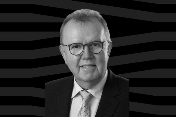 Ein Schwarz-Weiß-Porträt zeigt eine Person mit Brille in formeller Kleidung vor einem dunklen grafischen Hintergrund. Die Aufnahme zeigt Kopf, Schultern und den oberen Teil des Oberkörpers. Die Person trägt ein Sakko, ein gestreiftes Hemd und eine gemusterte Krawatte. Die Person blickt in Richtung Kamera. Das Bild ist als Freisteller gestaltet, sodass sich die dargestellte Person deutlich vom Hintergrund abhebt.
