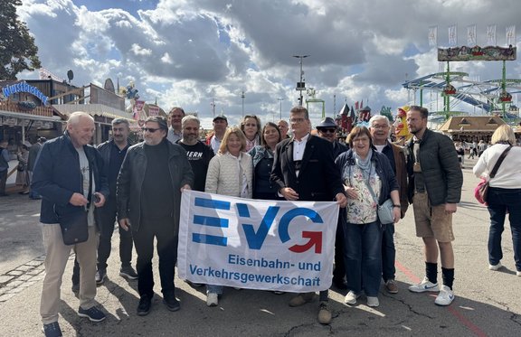 Gruppe von etwa 15 Personen auf einem Festgelände, einige halten ein weißes Banner mit dem blauen und roten Schriftzug „EVG“ sowie dem Text „Eisenbahn- und Verkehrsgewerkschaft“. Im Hintergrund sind Verkaufsstände, Fahrgeschäfte einer Achterbahn, weitere Besucherinnen und Besucher sowie ein bewölkter Himmel zu sehen.