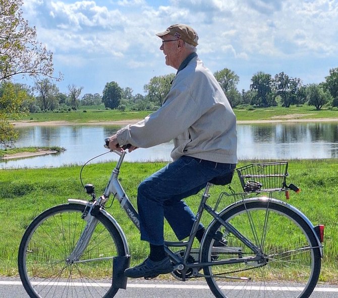 Eine einzelne Person fährt auf einem Fahrrad von links nach rechts auf einer Straße oder einem Weg. Im Vordergrund ist das Fahrrad mit Gepäckträger zu sehen. Die Person trägt eine Kappe, eine helle Jacke und dunkle Hosen. Im Hintergrund befindet sich eine grüne Landschaft mit Wiesen, einzelnen Bäumen und einem Gewässer. Der Himmel ist blau mit weißen Wolken.