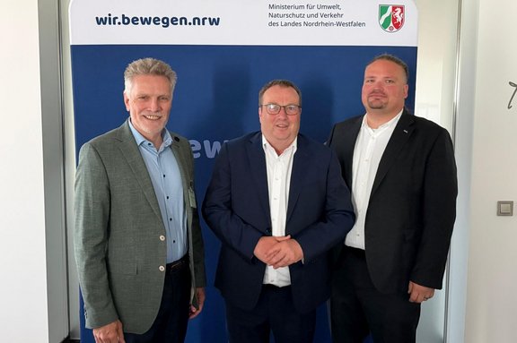 Drei Personen in Anzug beziehungsweise Sakko stehen nebeneinander vor einem Roll-up-Banner. Auf dem Banner stehen oben links „wir.bewegen.nrw“ und oben rechts „Ministerium für Umwelt, Naturschutz und Verkehr des Landes Nordrhein-Westfalen“ neben dem Wappen von Nordrhein-Westfalen. Der Hintergrund des Banners ist überwiegend blau. Links und rechts sind Teile einer hellen Innenraumwand sichtbar.