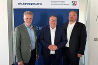 Drei Personen in Anzug beziehungsweise Sakko stehen nebeneinander vor einem Roll-up-Banner. Auf dem Banner stehen oben links „wir.bewegen.nrw“ und oben rechts „Ministerium für Umwelt, Naturschutz und Verkehr des Landes Nordrhein-Westfalen“ neben dem Wappen von Nordrhein-Westfalen. Der Hintergrund des Banners ist überwiegend blau. Links und rechts sind Teile einer hellen Innenraumwand sichtbar.